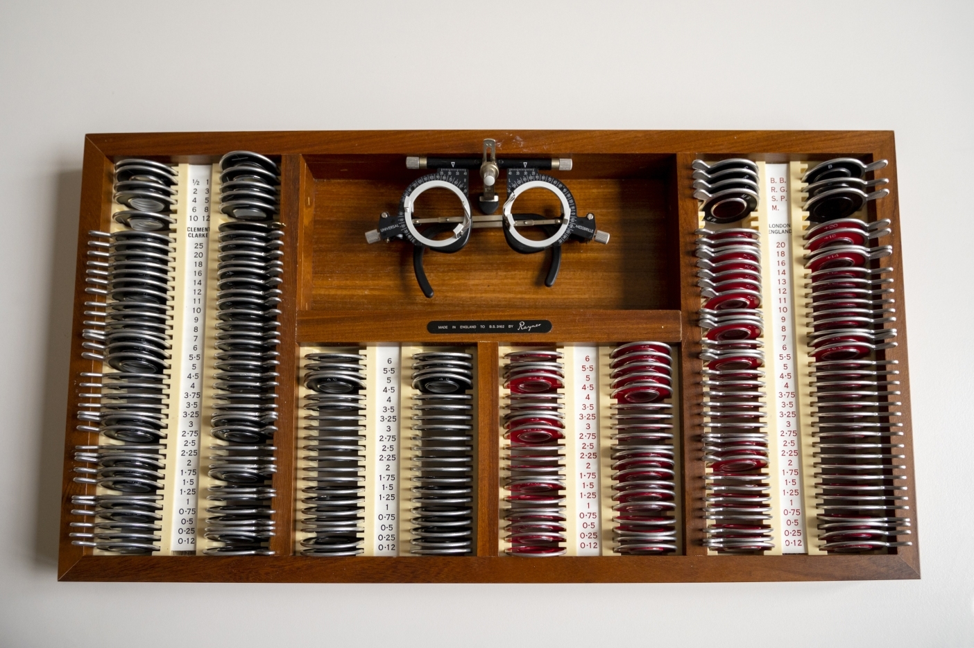 A close-up of an optical testing tool with a set of black and red lenses, along with a pair of eye test frames. The equipment is neatly organised in a wooden frame, with lens power details clearly marked next to each set.