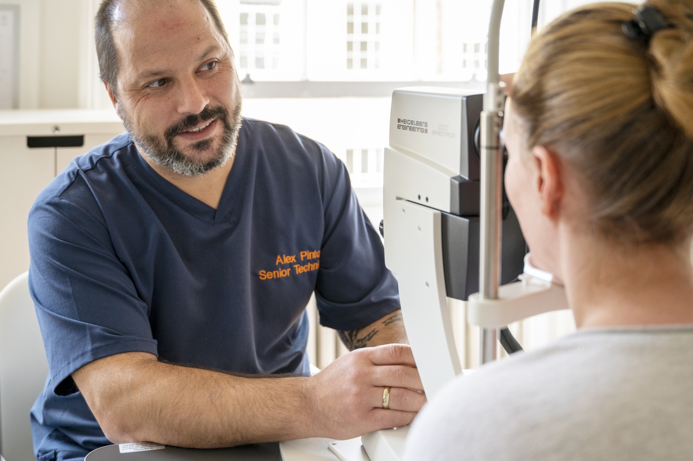 A male optometrist wearing a blue uniform and name tag is explaining an eye examination procedure to a female patient in an eye exam room. The optometrist is seated across from the patient, who is facing an eye-testing machine.